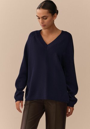 Dunkelblauer, übergroßer Pullover mit V-Ausschnitt, gerippten Akzenten und langen Ärmeln, kombiniert mit dunkelbraunen Lederhosen. Weiche Textur.