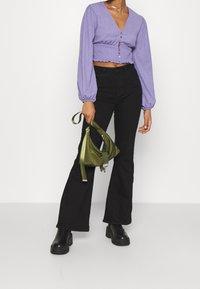 Lila, kurz geschnittene Bluse mit geraffter Textur, langen Puffärmeln, schwarze ausgestellte Jeans und eine grüne Tasche mit verstellbarem Riemen. Schwarze Plateaustiefel.