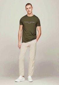 Olivgrön bomulls-T-shirt med vit text "TOMMY HILFIGER EST. 1985," kombinerad med beige byxor och vita sneakers. Enkel, figurnära design.