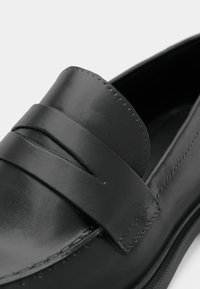 Black leather loafer with a smooth finish, featuring a wide strap across the front and detailed stitching along the edges.