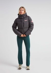 Sort pufferjakke med høj krave, quiltet design og pink logo detaljer. Sammen med grønne leggings og lyse sko.