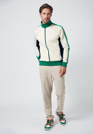 Veste de sport crème avec des accents verts et navy, col haut, fermeture éclair à l'avant ; pantalon beige ; baskets vertes avec lacets orange et détails.