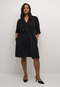 Robe noire à ceinture, fabriquée en tissu texturé ; présente un décolleté en V, des manches roulées et des poches latérales. Portée avec des chaussures à talons hauts noires.