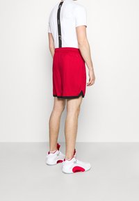 Hombre vestido con una camiseta blanca, pantalones cortos deportivos rojos con ribete negro y zapatillas de baloncesto blanco y rojo, de pie contra un fondo claro y liso.