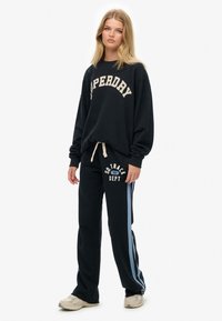 Granatowy sweatshirt z białym logo „Superdry”, w połączeniu z czarnymi dresami z białymi i jasnoniebieskimi akcentami oraz napisem. Wygodny materiał i luźny krój.