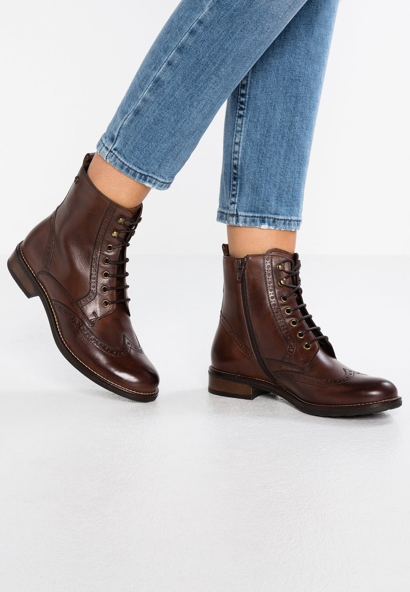 Personne portant un jean bleu et des bottines en cuir marron à lacets avec des fermetures éclair latérales, debout sur un sol blanc.