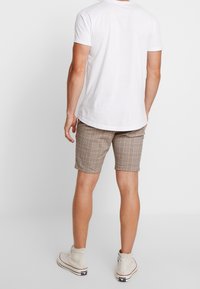 Pánský casual outfit, který zahrnuje bílé tričko a béžové kostičkované kraťasy. Kraťasy mají kostkovaný vzor a jsou kombinovány se světlými botami.