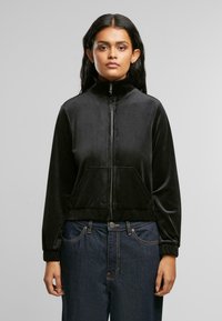 Svart glidelåsvelourjakke med høy krage, lange ermer og frontlommer. Har elastiske mansjetter og hem, brukt over blå denimjeans.