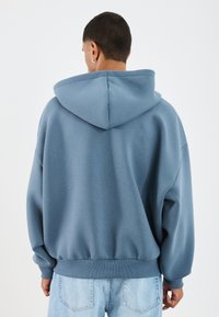 Blauw hoodie met een ontspannen pasvorm, ribbelboorden aan de onderkant en mouwen. Heeft een grote capuchon en een zacht, textuurstof. Achteraan gezien, gecombineerd met lichte jeans.