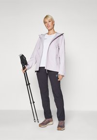 Šviesiai violetinė vandeniui atspari striukė su kapišonu, po ja balta marškinėliai, tamsios kelnės ir rudos žygių bateliai; laikanti juodus žygių lazdas.