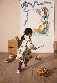 Kind in einem gemusterten Kleid mit Puffärmeln, das einen Pinsel hält, steht auf einem Holzboden neben bunten Spielzeugen und einem dekorierten Wandgemälde.