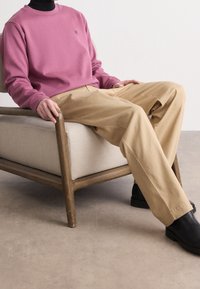 Felpa rosa e pantaloni beige indossati mentre si è seduti su una poltrona rivestita in tessuto chiaro con braccioli in legno, abbinati a scarpe nere.