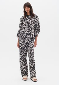 Femeie îmbrăcată cu un top larg cu print abstract alb și negru și pantaloni evazați asortați, purtând sandale bej cu talpă joasă, pe un fundal alb.