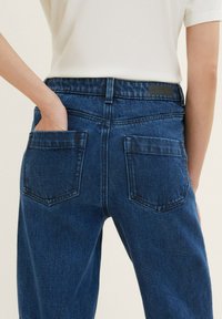Personne portant un jean en denim bleu foncé avec des poches arrière, une main partiellement dans la poche gauche, et un haut blanc côtelé.