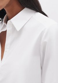 Camicia bianca con colletto, caratterizzata da un tessuto testurizzato e un design semplice. Primo piano dei dettagli della camicia, con pulsanti e manica.