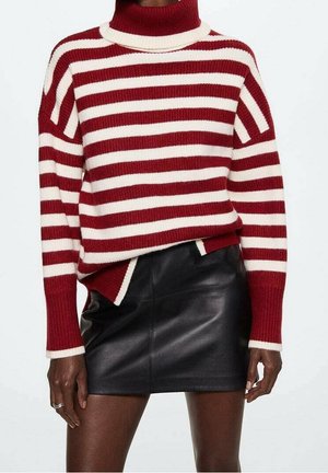 Femme portant un pull à col roulé rayé rouge et blanc et une mini-jupe en cuir noire, debout devant un fond uni.