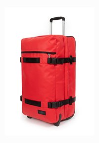Valise en tissu rouge de forme rectangulaire, dotée de sangles noires, d'une poche frontale zippée et d'une poignée rétractable.