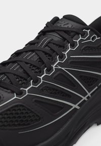 Κοντινό πλάνο μαύρου αθλητικού παπουτσιού με περίπλοκα γκρι γεωμετρικά σχέδια και μαύρα κορδόνια με λευκή ραφή στη μέση.