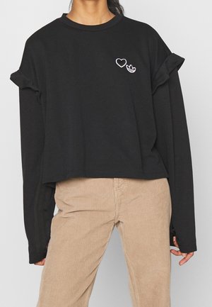 Kobieta ubrana w czarną krótka bluzę z długim rękawem ozdobioną naszywkami w kształcie serca i logo, w połączeniu z beżowymi spodniami ze sztruksu.