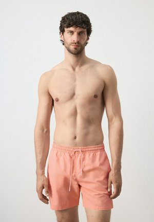 Giovane uomo con capelli ricci scuri che indossa pantaloncini da bagno color corallo, in piedi contro uno sfondo chiaro e uniforme, rivolto verso la fotocamera.