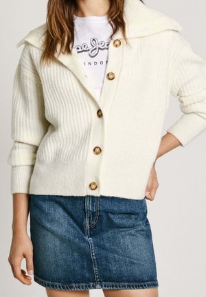 Vrouw die een crème ribgebreide cardigan met knopen draagt over een wit T-shirt met een print, en een blauwe spijkerrok, met één hand op haar heup.