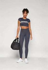 Mulher em top cropped azul-marinho e leggings segurando uma bolsa de ginásio preta, de pé sobre um fundo branco e usando tênis brancos.