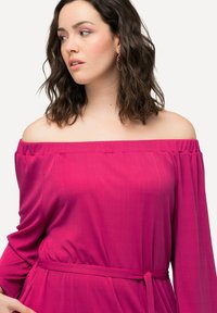 Olkapäiltä avonainen kirkkaan vaaleanpunainen paita, jossa on ribbikuviointi, pitkät hihat ja kiristettävä vyötärö, johon kuuluu samanvärinen kangasvyö.