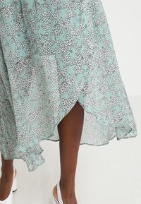 Een vloeiende mintgroene jurk met een bloemenpatroon, voorzien van een gerimpelde zoom en een split aan één zijde, gemaakt van lichtgewicht stof.