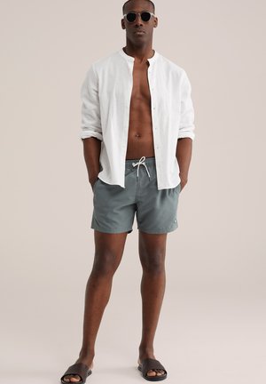 Egy férfi kerek napszemüveget visel, nyitott fehér gombos inget, szürke fürdőnadrágot és sötét szövött szandált, semleges háttér előtt áll.