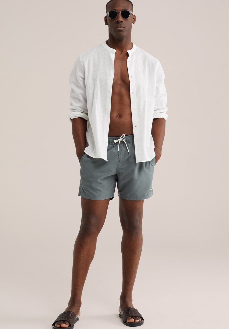 Homme portant des lunettes de soleil rondes, une chemise blanche à boutons ouverte, un short de bain gris et des sandales tissées foncées, debout devant un arrière-plan neutre.