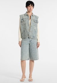 Jasnoniebieska kamizelka dżinsowa bez rękawów z kieszeniami z przodu, w komplecie z dopasowanymi szortami do kolan i białymi klapkami.