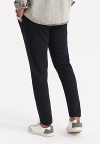 Zwarte taps toelopende broek van een gestructureerde stof, gecombineerd met witte sneakers met een grijze accent. Zicht van achter.