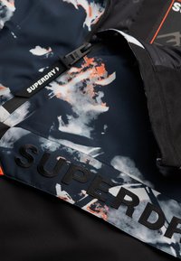 Black and navy patterned jacket featuring graphic prints in orange and gray, with prominent "SUPERDRY" branding and textured fabric accents.