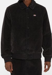 Czarna kurtka z korduroju z kołnierzykiem, zapięciem na guziki i elastycznymi mankietami. Posiada małą naszywkę z logo na lewej piersi oraz boczne kieszenie.