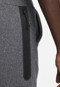 Šedé teplákové kalhoty z látky s černým zipovým kapsou, ozdobené texturovaným poutkem a hladkým jazýčkem, které zdůrazňují moderní design a funkčnost.