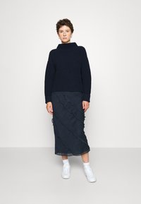 Maglione a coste blu navy con colletto alto, abbinato a una gonna di media lunghezza blu scuro con dettaglio di tessuto drappeggiato. Sneakers bianche completano l'outfit.