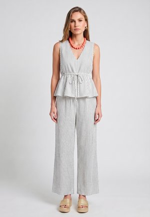 Donna che indossa un top bianco senza maniche a righe con cintura in vita, pantaloni larghi abbinati, sandali beige con zeppa e collana di perline corallo su sfondo neutro.