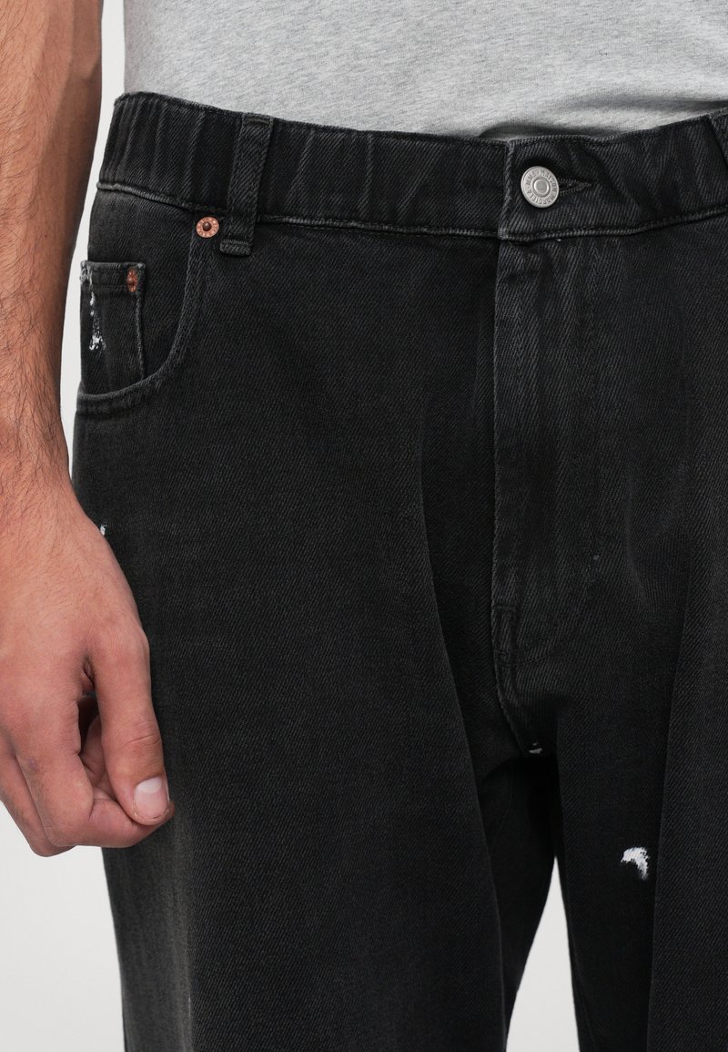 Black denim jeans featuring a relaxed fit, elastic waistband, and distressed detailing on pockets. Sleek texture and minimalistic design.
