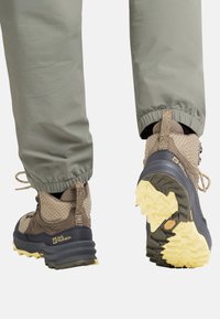 Scarponcini da trekking marroni e blu con una tomaia texture, caratterizzati da una rete decorata e una suola gialla spessa. I pantaloni sono infilati nei polsini.
