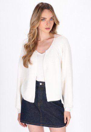 Vrouw met lang blond haar die een witte cardigan over een wit topje en een donkerblauwe spijkerrok draagt, staand voor een effen achtergrond.
