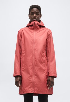 Άτομο που φοράει ροδακινί κόκκινο αδιάβροχο με κουκούλα και εμπρόσθιο φερμουάρ, στέκεται μπροστά σε απλό ανοιχτό γκρι φόντο.