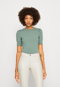 Top se širokými rukávy, přiléhavý, ve světlé zelené látce, doplněný bledě béžovými skinny džíny. Má kulatý výstřih a hladkou strukturu.