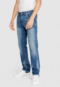 Jeans de mezclilla azul con corte recto, ligero desgastado, cinco bolsillos y botón de tono cobre. Combinados con zapatillas blancas y una camiseta básica.