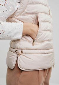 Jasnoróżowa kamizelka puchowa z poziomym pikowaniem, boczna kieszeń z złotym suwakiem, regulowany sznurek u dołu, nałożona na białą bluzkę z ażurowym wzorem.