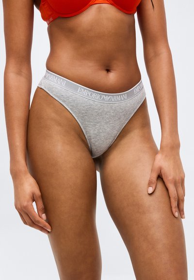 Γυναίκα που φοράει γκρι εσώρουχα μπικίνι Emporio Armani και πορτοκαλί σουτιέν, όρθια με τα χέρια χαλαρά στο πλάι, μπροστά σε απλό φόντο.