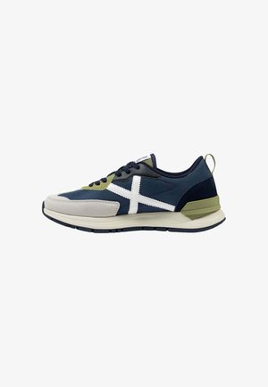 Zapatilla deportiva azul marino y oliva con logo X blanco lateral, cordones negros, suela beige y acento verde en el talón, vista lateral.