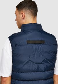 Navyblauwe puffer vest, met een hoge kraag, gestikt ontwerp en een opvallende bladeren borduurwerk op de achterkant. Glad, synthetisch materiaal.