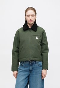 Groene jas met zwarte, fuzzy kraag, ritssluiting, zijzakken en een klein logo-embleem. Gecombineerd met blauwe wijde denim jeans.