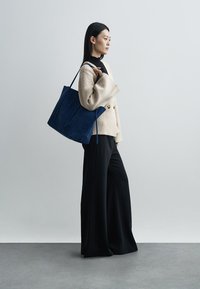 Blue suede tote bag with long handles, positioned next to a model wearing a beige textured blazer and black wide-leg pants, against a gray backdrop.