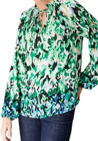 Blouse à manches longues avec imprimé abstrait vert, noir, bleu et blanc, encolure froncée avec cordon, portée avec un jean bleu foncé.